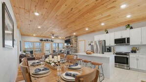 A spacious dining area with a wooden table set for four, surrounded by wooden chairs. A stone fireplace and a large window with a view of trees are visible in the background. - Modern Smoky Mountain Home w/ Hot Tub, Game Room, Theater, Sleeps 12 (Sevierville)