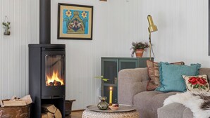 Un salon chaleureux avec un poêle à bois, un canapé confortable, une table basse ronde en osier et une œuvre d’art encadrée accrochée au mur.
