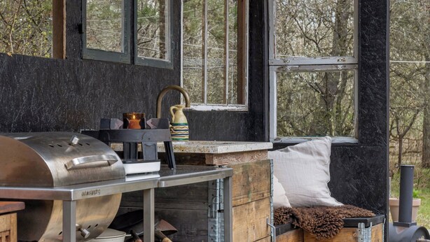 L’intérieur d’un chalet rustique, avec un poêle à bois, un gril en métal et une fenêtre donnant sur une forêt.