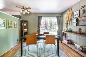 Un coin repas comprenant une table, des chaises et un ventilateur de plafond. Les murs sont ornés de tableaux encadrés et il y a une fenêtre avec des rideaux.