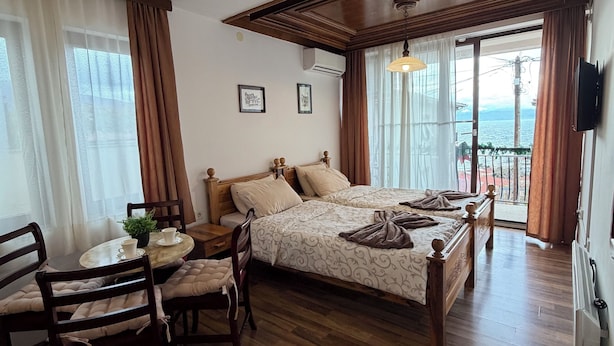 Un dormitorio con suelo de madera, una cama con cabecera, una mesita de noche con una planta y una ventana con cortinas.
