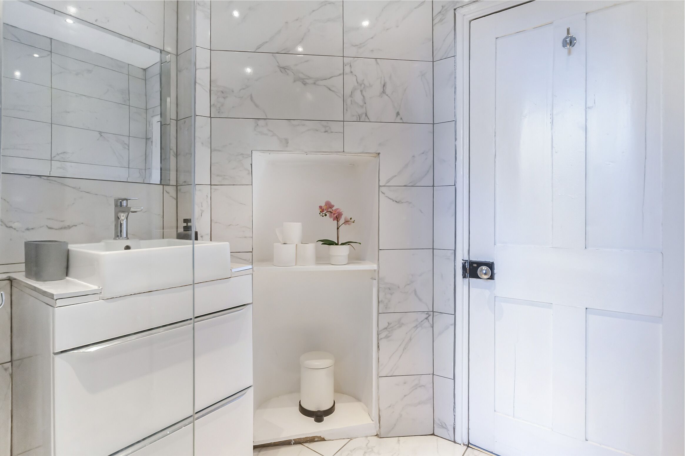 A modern bathroom with a marble-tiled wall, a white sink, and a shower area with a small shelf holding toiletries.