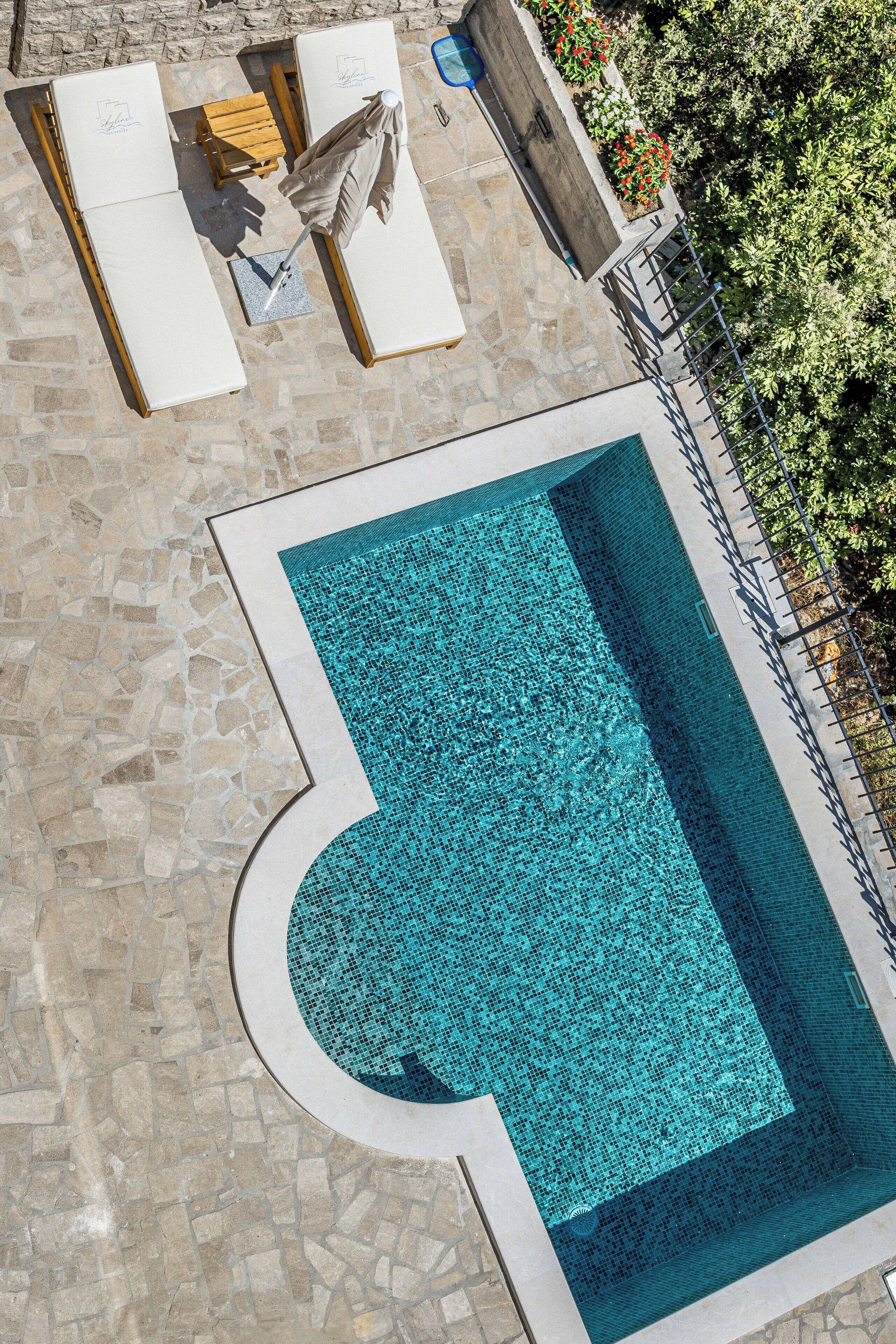 A swimming pool with a tiled deck, lounge chairs, and a wooden table.