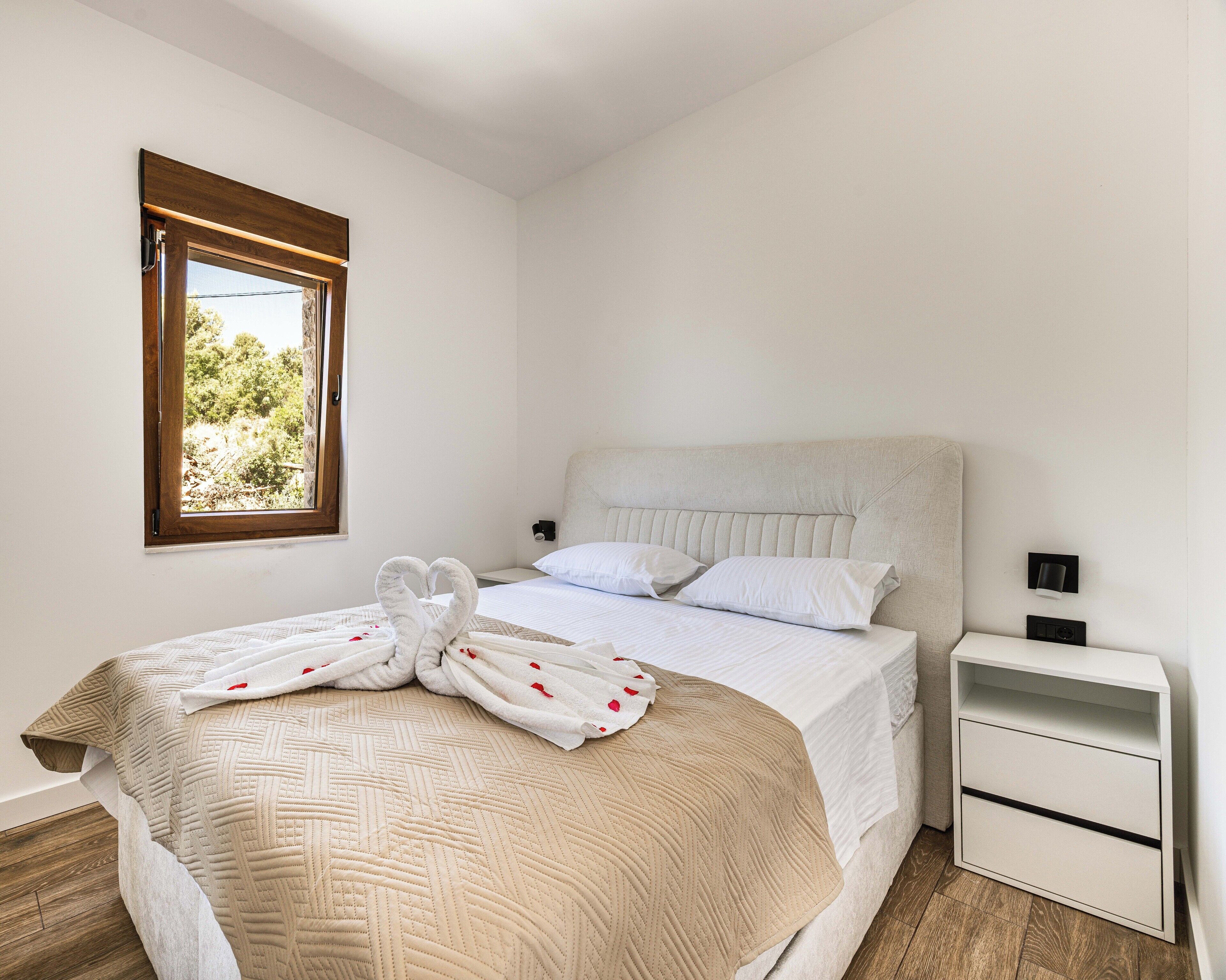 Un lit bien fait, recouvert d’une courtepointe beige, avec deux oreillers blancs et une serviette en forme de cygne posée sur le lit. On y trouve une petite table de chevet avec une lampe et un tiroir.