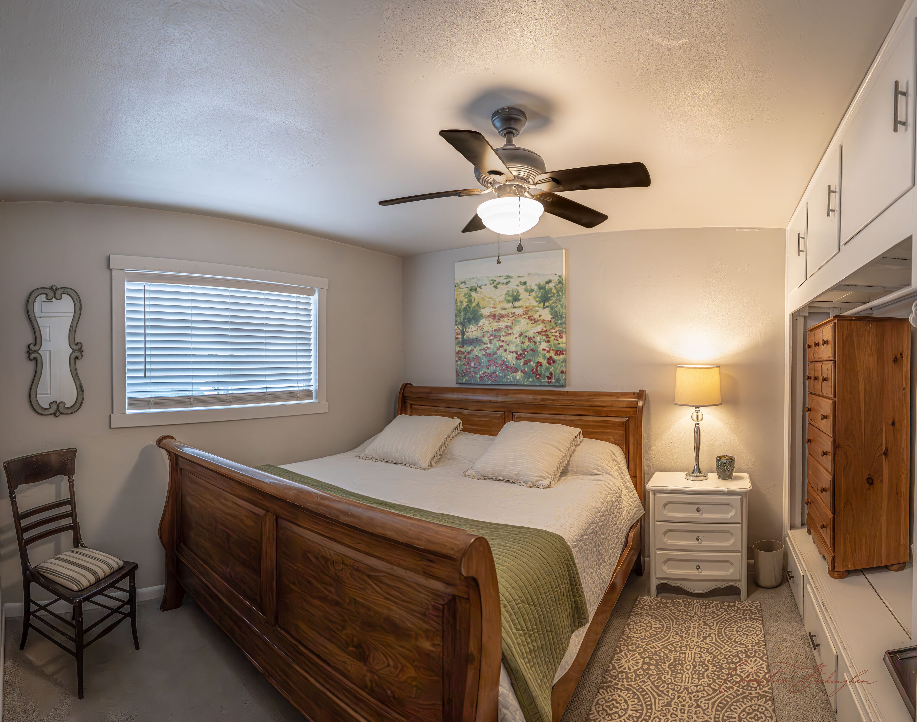 A bedroom with a wooden bed, a ceiling fan, a bedside lamp, a window with blinds, a chair, a dresser, and a painting on the wall.