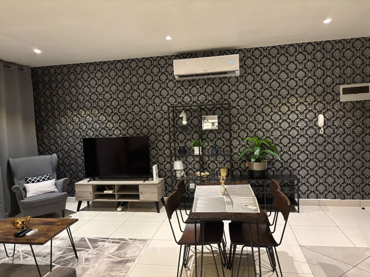 A modern living room with a flat-screen TV, a grey armchair, a dining table with chairs, and a patterned wallpaper.