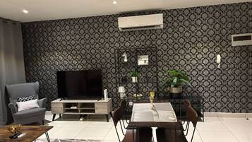 A modern living room with a flat-screen TV, a grey armchair, a dining table with chairs, and a patterned wallpaper.