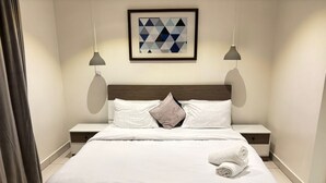 A neatly made bed with white linens and a rolled towel on the side. There are two bedside tables with lamps, a framed geometric artwork on the wall, and a window with curtains. - Embassy Onebedroom by Unique Abodes (Accra)