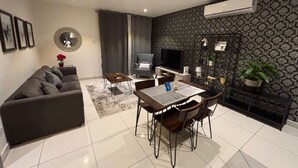 A modern living room with a sofa, dining table, and chairs. There is a TV, a plant, and a patterned wall. - Embassy Onebedroom by Unique Abodes (Accra)