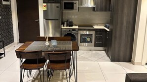 A modern kitchen with dark cabinetry, stainless steel appliances, and a dining area with a glass table and wooden chairs. - Embassy Onebedroom by Unique Abodes (Accra)