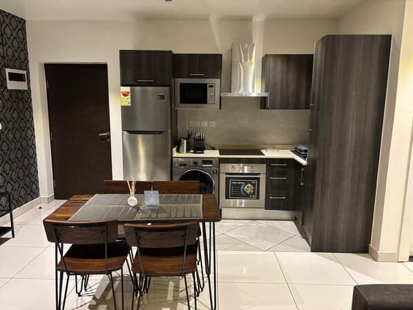 A modern kitchen with dark cabinetry, stainless steel appliances, and a dining area with a glass table and wooden chairs. - Embassy Onebedroom by Unique Abodes (Accra)