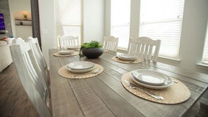 Een eettafel gedekt met borden, bestek en placemats in een lichte kamer met witte muren en grote ramen.