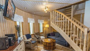 Une pièce chaleureuse avec des murs en bois, un canapé, deux fauteuils, une table basse, un escalier en bois et un téléviseur fixé au mur.