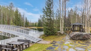 Une terrasse en bois avec une rambarde en métal, donnant sur un étang, entourée d’arbres et bordée d’un sentier en pierre.