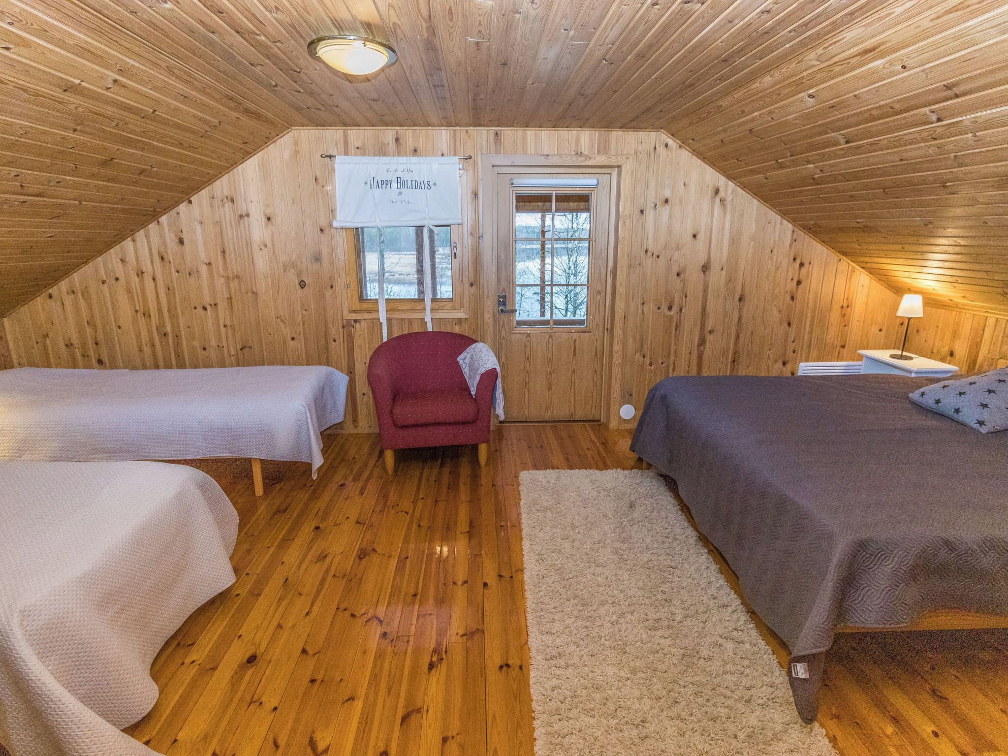A cozy attic bedroom with wooden walls, two single beds, a red armchair, and a window with a view of trees.