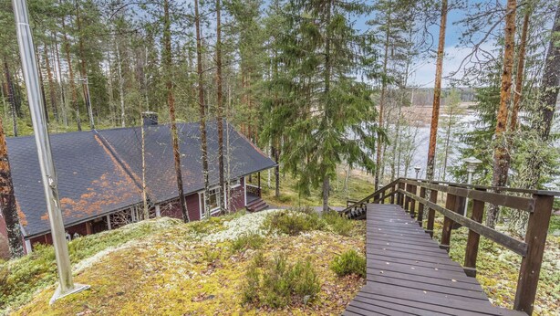 Une cabane dotée d’une terrasse en bois qui mène à un porche, entourée de grands pins et avec une étendue d’eau en arrière-plan.