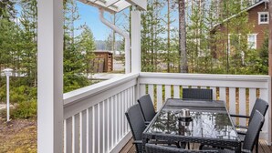 Een overdekt terras buiten met een tafel met glazen blad en rieten stoelen, omgeven door een witte balustrade en uitzicht op een bebost gebied.