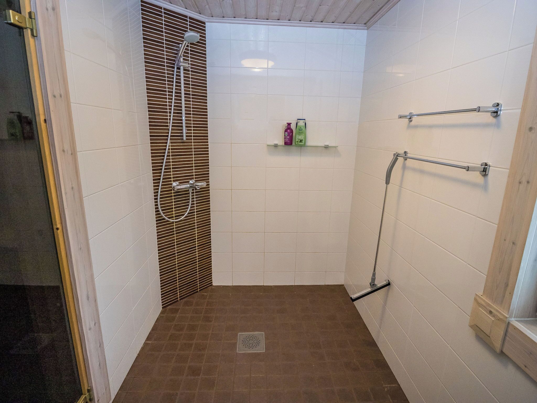 A small, enclosed shower area with a showerhead and a handheld shower attachment. There is a shelf with toiletries and a towel rack on the wall.