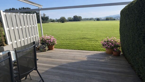 Een buitenterras met houten terras, een metalen pergola en uitzicht op een groen veld.