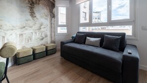 Un soggiorno moderno con un divano grigio scuro, una poltrona verde e un poggiapiedi rotondo. Si vede una grande finestra con vista sugli edifici e un murale sulla parete.