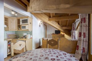 Ein kleines Zimmer mit Bett, einer Küchenzeile und einem schmales Fenster mit Vorhängen.