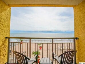 Uma varanda com vista para a praia, duas cadeiras e uma mesa pequena.
