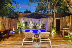 Un patio exterior con terraza de madera, dos sillas de madera, una mesa, un paraguas grande y una piscina pequeña rodeada de plantas.