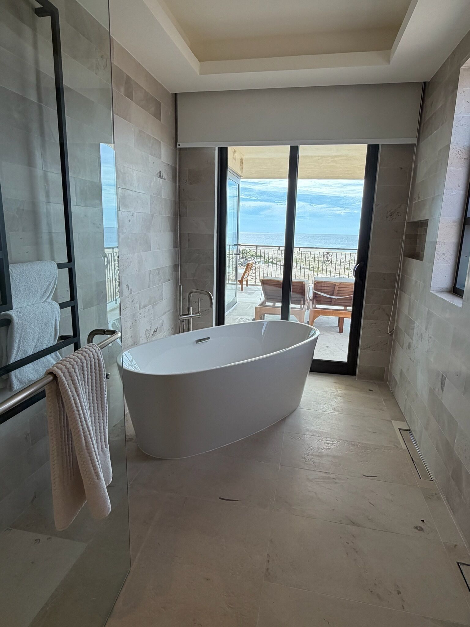 A modern bathroom with a freestanding bathtub, a glass shower enclosure, and a view of a balcony with outdoor furniture.