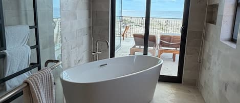 A modern bathroom with a freestanding bathtub, a glass shower enclosure, and a view of a balcony with outdoor furniture.