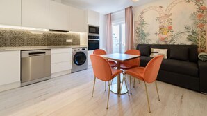 Eine moderne Küche mit weißen Schränken, einem Geschirrspüler und einer Waschmaschine. Ein Essbereich mit Tisch und orangefarbenen Stühlen ist ebenfalls vorhanden.
