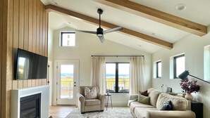 A spacious living room with a high ceiling, a fireplace, a television mounted on the wall, a sofa, a chair, and a view of the countryside through large windows. - Sunset Views & Ranch Life ~ Targhee Barndominium (Alta)