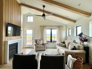 A spacious living room with a high ceiling, a fireplace, a television mounted on the wall, a sofa, a chair, and a view of the countryside through large windows. - Sunset Views & Ranch Life ~ Targhee Barndominium (Alta)