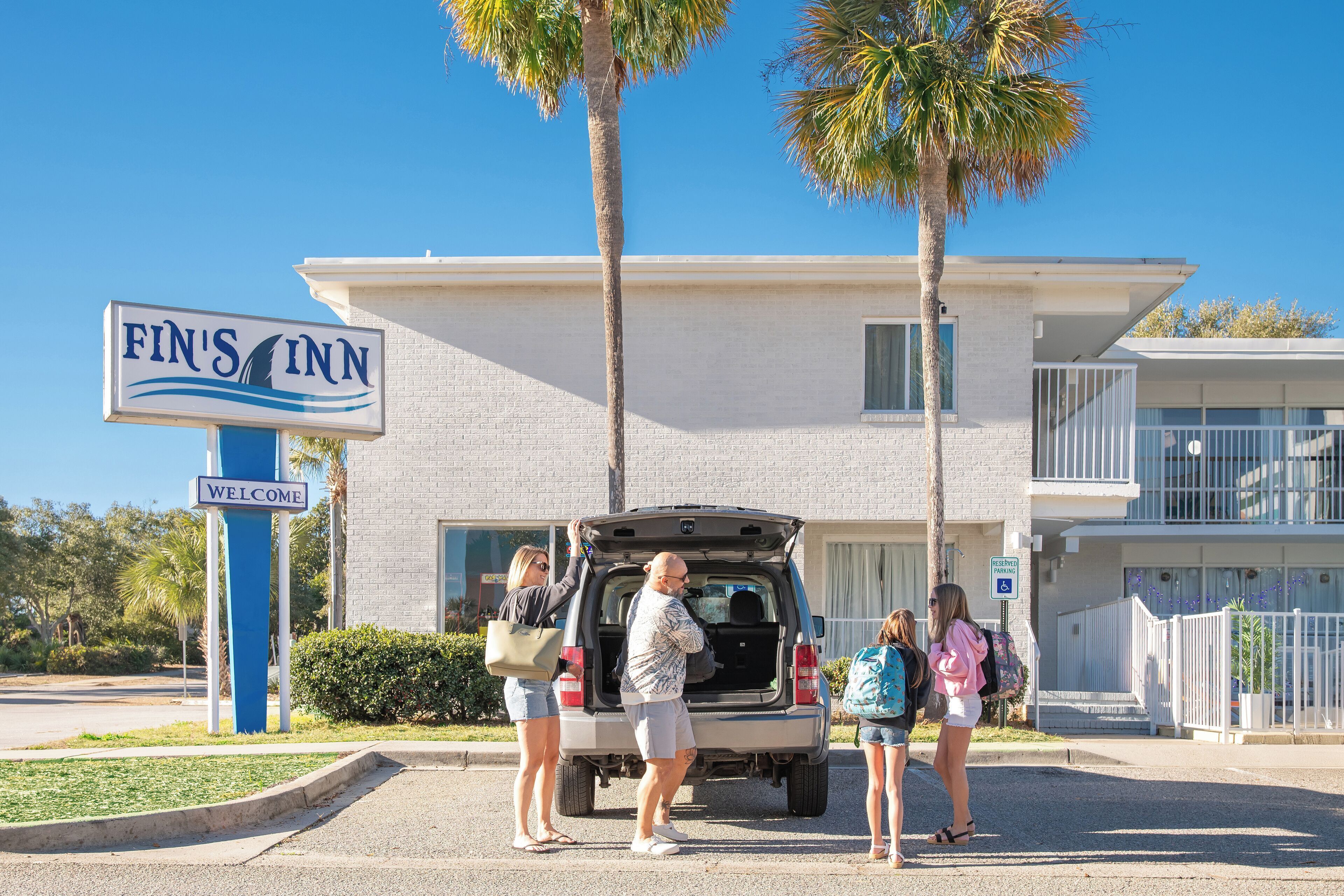 A Fin's Inn sign with a welcome message, a building with a white facade, and a parking area with people and a vehicle.