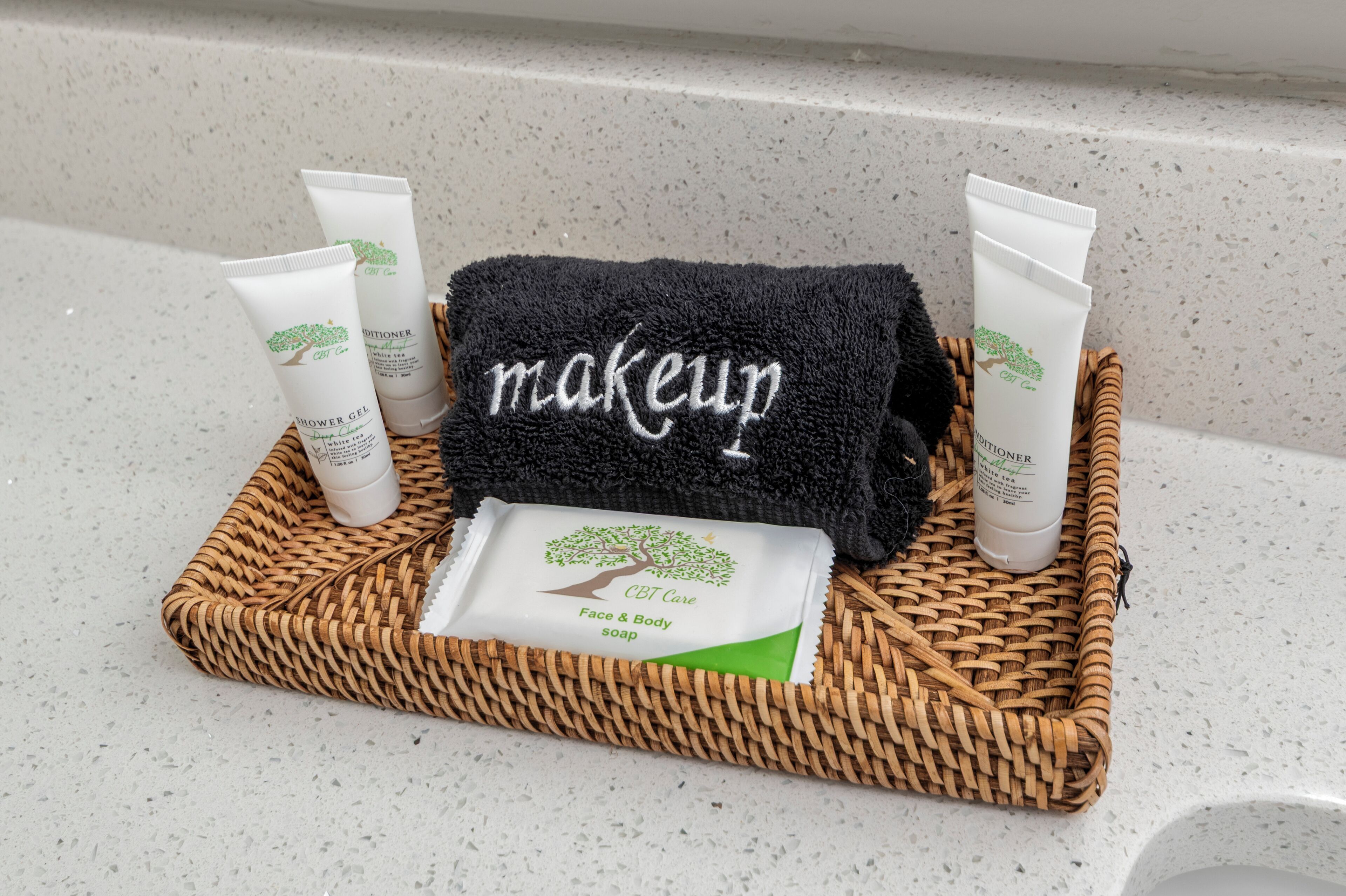 A bathroom counter with a woven basket containing a black towel labeled "makeup," a face and body soap, and two tubes of lotion.