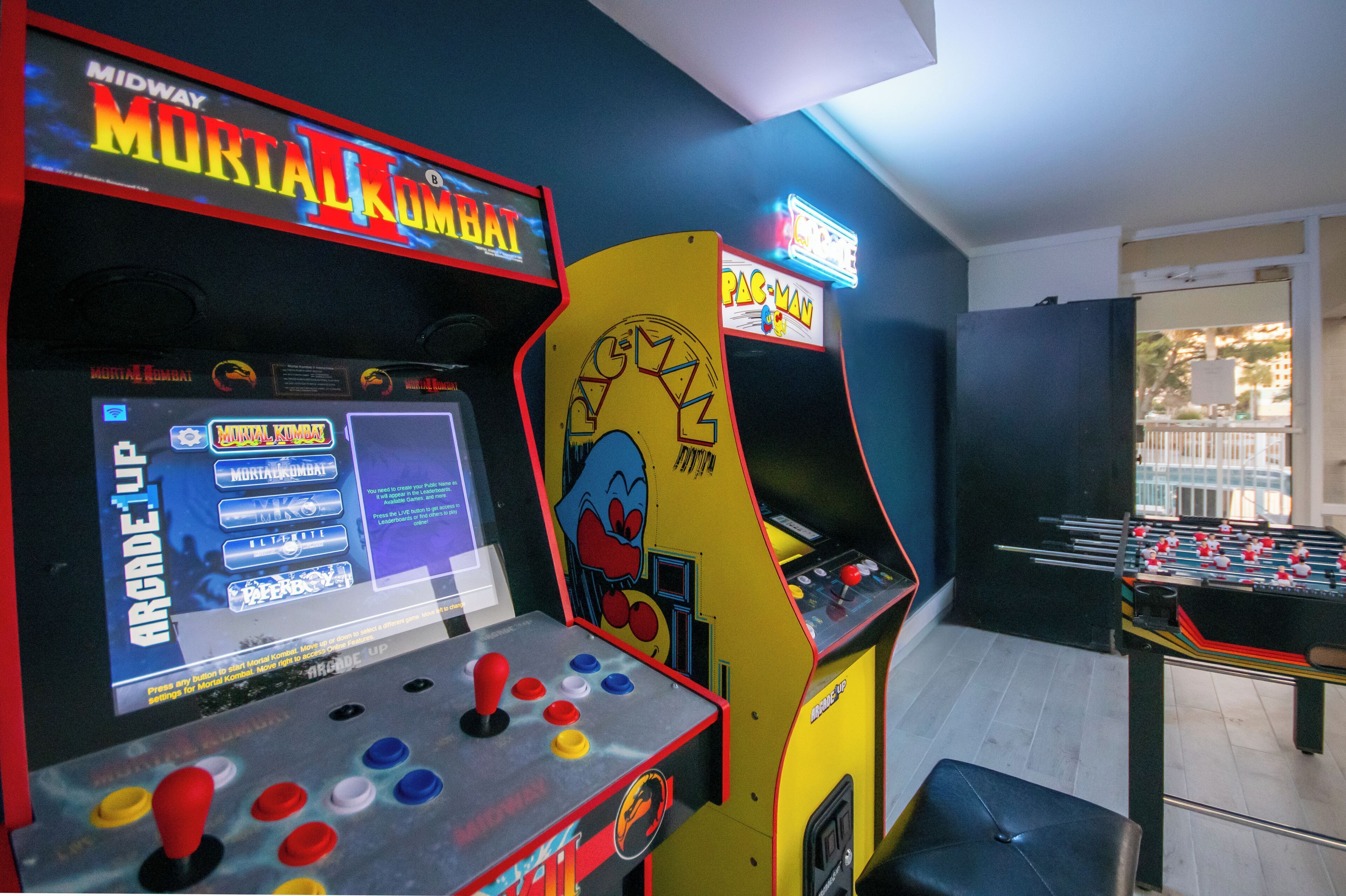 Arcade machines including Mortal Kombat and Pac-Man, a foosball table, and a window with a view of the outside.