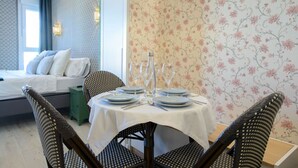 Ein Essbereich mit einem runden Tisch, gedeckt für vier Personen, umgeben von Korbstühlen. Im Hintergrund ist ein Bett mit weißen Leinen zu sehen, und der Raum ist mit blumigem Wandpapier gestaltet.
