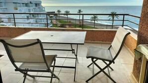 A balcony with a table and two chairs, overlooking a beach and a white building.