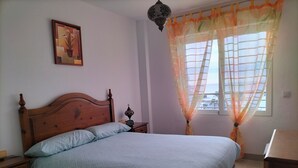 A bedroom with a wooden bed, a nightstand, a bedside table, a wall-mounted clock, and a window with sheer curtains.