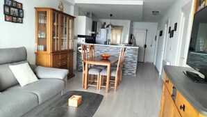A modern living room with a grey sofa, a dining table with chairs, a wooden cabinet, and a kitchen in the background.