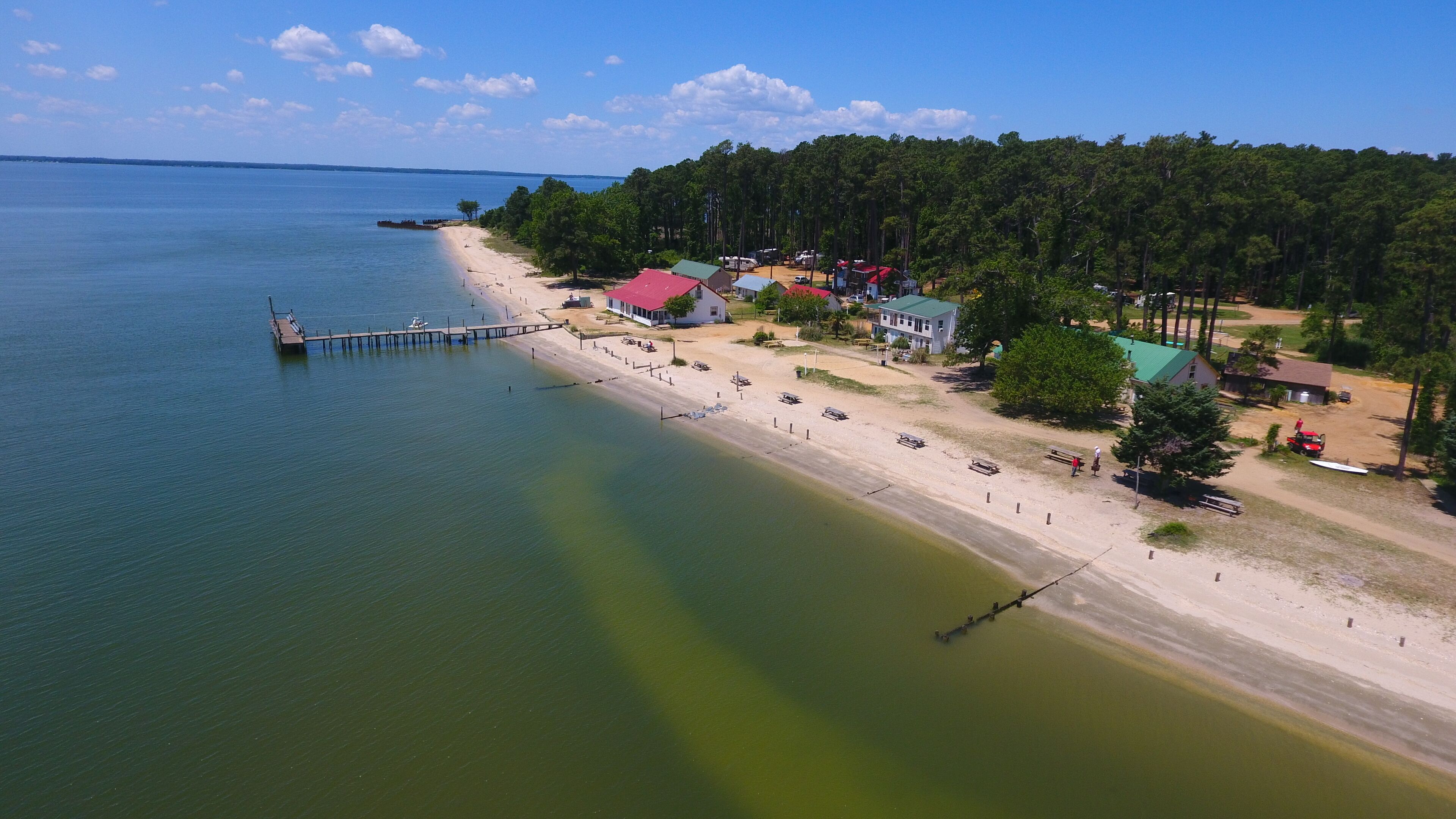An aerial view of a coastal area with a pier extending into the water, a sandy beach, and several buildings with red roofs.