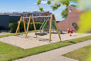 A playground with swings and a slide, surrounded by a brick building and greenery.