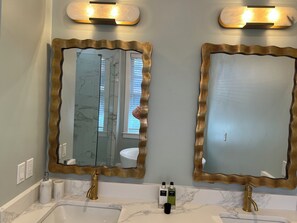 Ett badrum med två handfat, bänkskivor i marmor, speglar med guldfärgade ramar och väggmonterade lampetter.