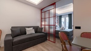 Quarto moderno e compacto, com sofá cinza, mesa de jantar e porta deslizante com moldura vermelha que dá acesso a um quarto com cama e vista para o exterior.