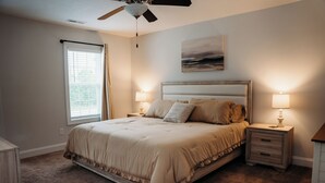Um quarto com uma cama grande, duas mesas de cabeceira com luminárias, um ventilador de teto, uma janela com cortinas e um quadro na parede.