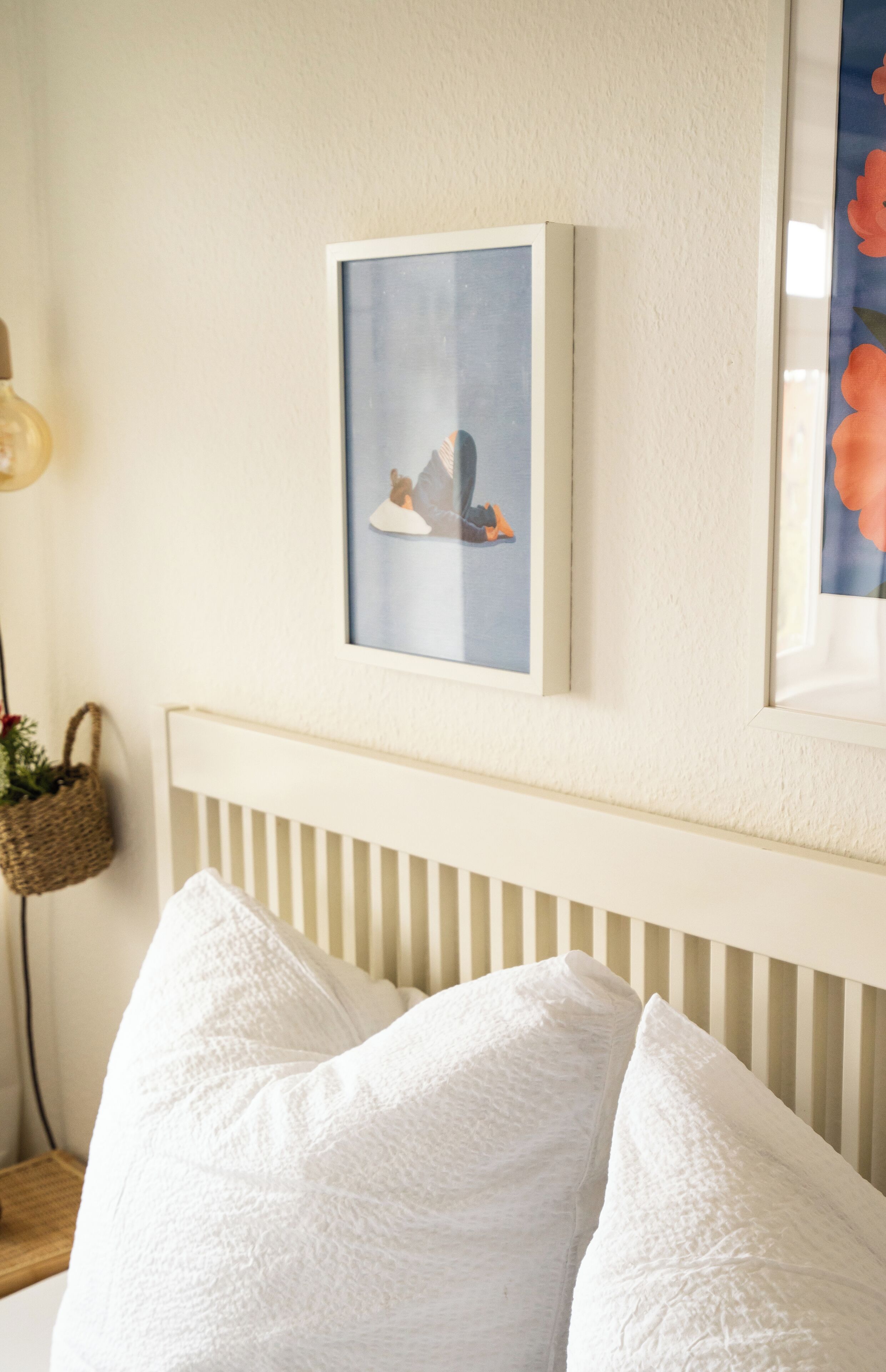 Une chambre à coucher avec une tête de lit blanche, un tableau encadré au mur et une table de chevet sur laquelle se trouvent une lampe et un panier.