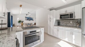 Una cucina moderna con mobili bianchi, un piano di lavoro in granito, elettrodomestici in acciaio inossidabile e un'area pranzo con una sedia blu e una pianta.