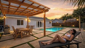 Un giardino con piscina, un pergolato in legno, un tavolo da pranzo con sedie e una poltrona da relax.