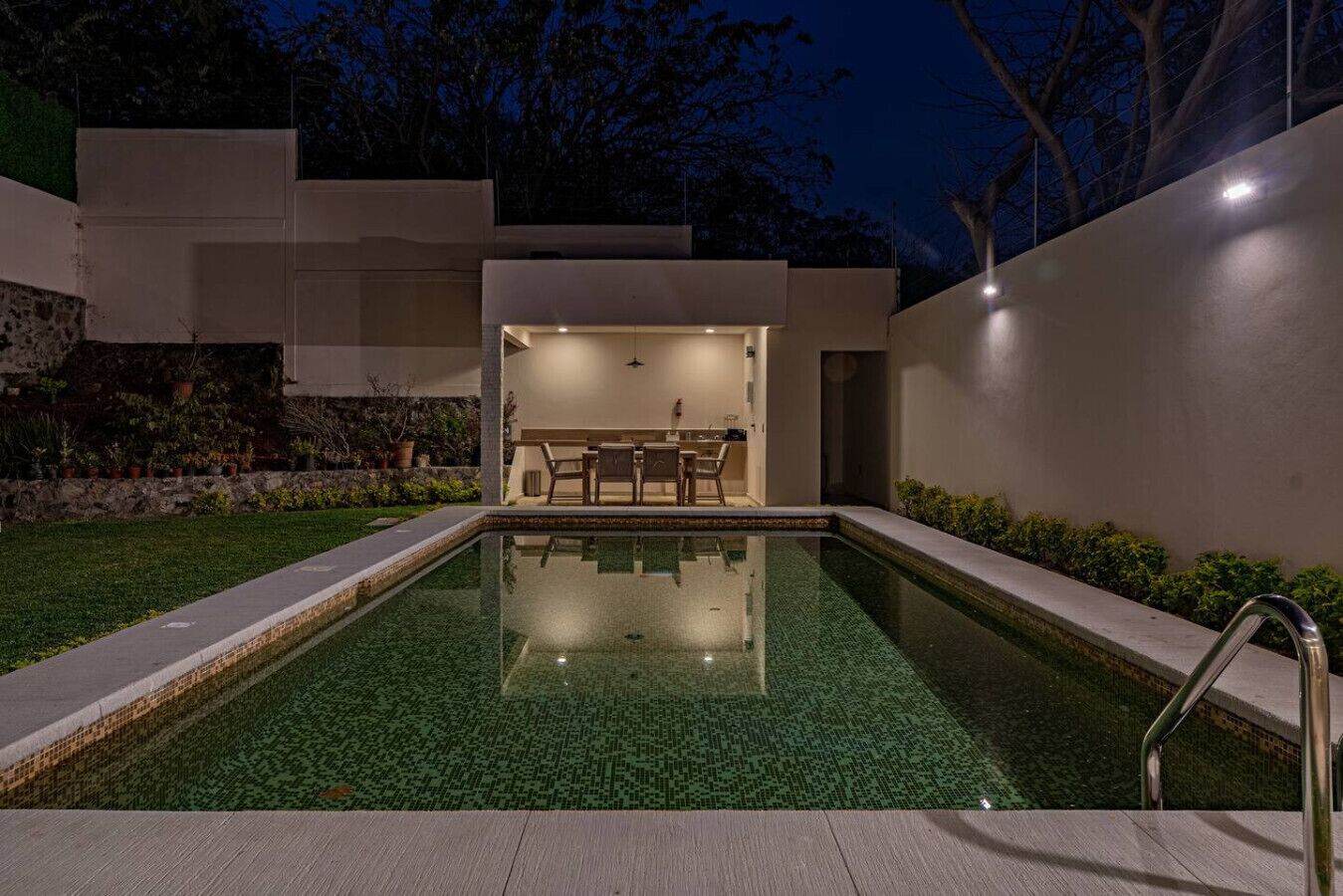 A modern outdoor pool area with a clear pool, a dining area with a table and chairs, and a stone wall with plants.