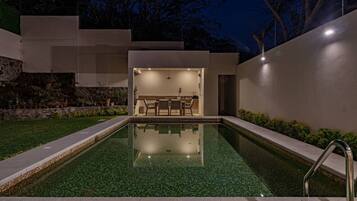 A modern outdoor pool area with a clear pool, a dining area with a table and chairs, and a stone wall with plants.