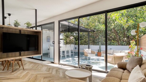 Un salon moderne doté d’un téléviseur, d’un canapé et d’un coin repas, de grandes portes-fenêtres coulissantes donnant sur une terrasse aménagée avec du mobilier d’extérieur et de la verdure.